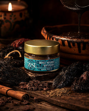 A ZEST jar of Mole Negro Mystery spice blend is centered on a dark wooden surface, surrounded by dried chiles, cocoa nibs, cumin, and a cinnamon stick, with a dark sauce pouring into a clay pot and a lit candle in the background.