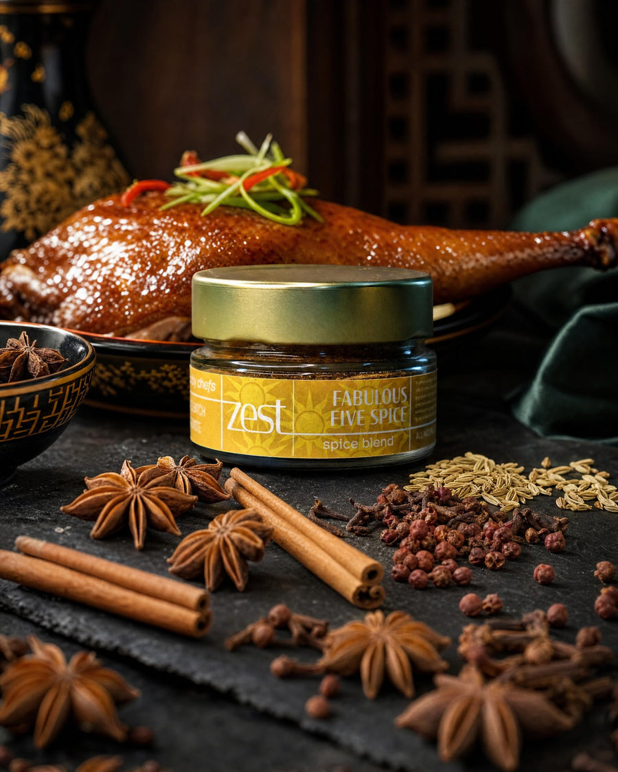 A jar of ZEST Fabulous Five Spice blend sits on a dark slate surface, surrounded by star anise, cinnamon sticks, cloves, Sichuan peppercorns, and fennel seeds, with a beautifully roasted duck in the background.