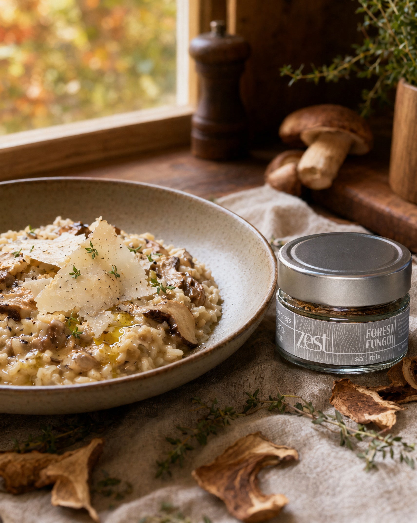 A bowl of mushroom risotto with parmesan and thyme, next to a ZEST Forest Funghi salt mix jar, fresh and dried mushrooms, and a pepper mill on a linen cloth and wooden table by a window.