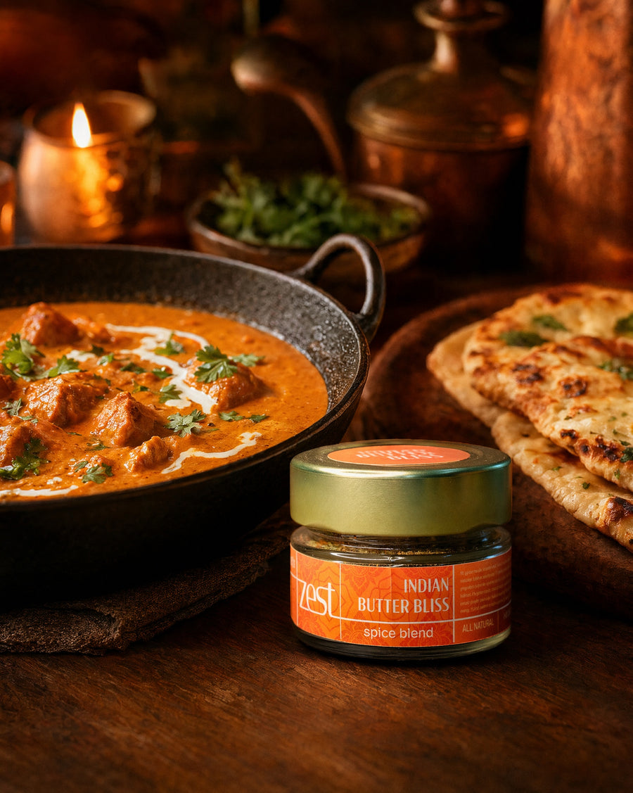 A ZEST Indian Butter Bliss spice blend jar displayed next to a rich butter chicken or paneer dish and naan bread on a dark wooden table, with candlelight in the background.