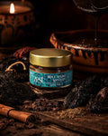 A ZEST jar of Mole Negro Mystery spice blend is centered on a dark wooden surface, surrounded by dried chiles, cocoa nibs, cumin, and a cinnamon stick, with a dark sauce pouring into a clay pot and a lit candle in the background.