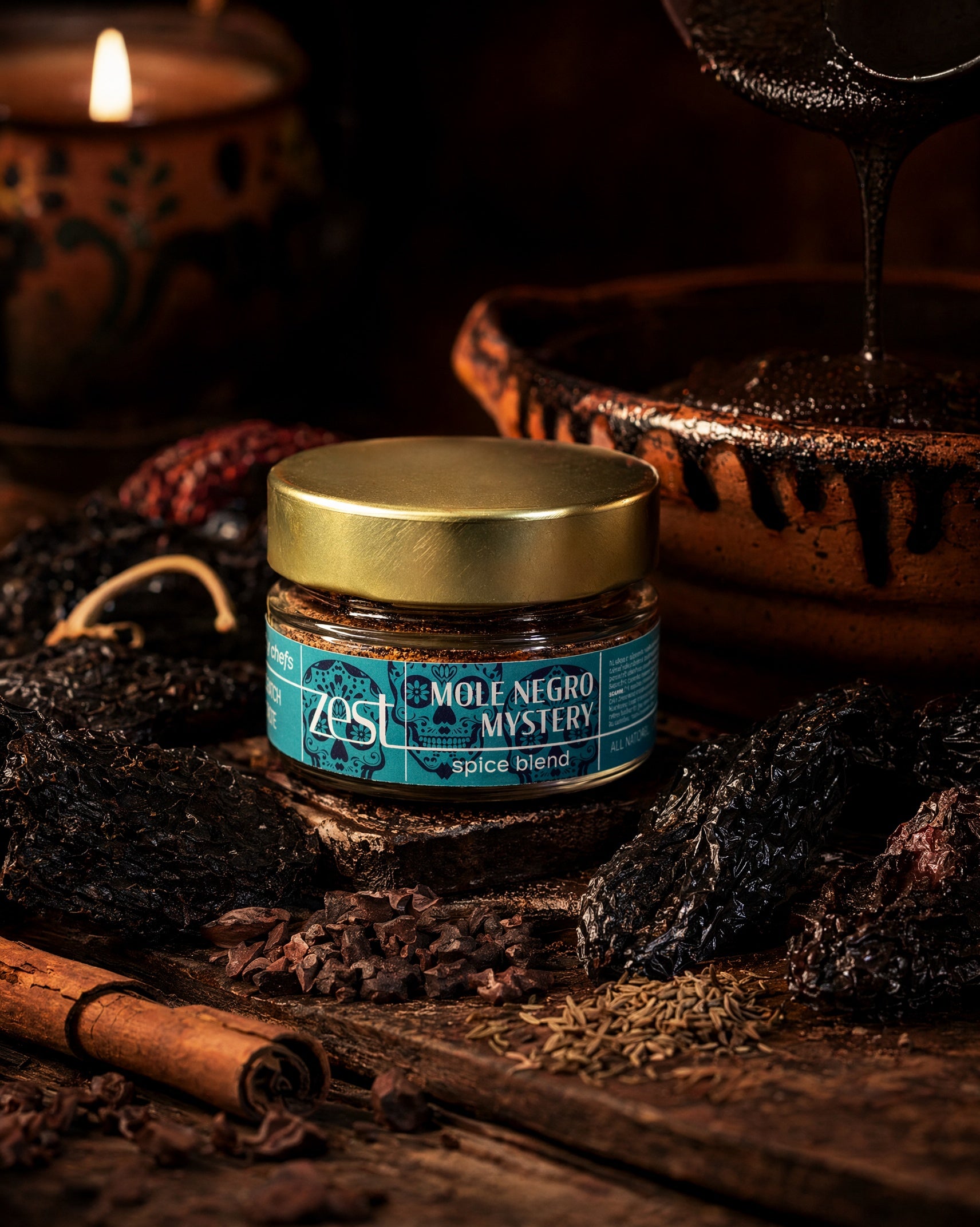 A ZEST jar of Mole Negro Mystery spice blend is centered on a dark wooden surface, surrounded by dried chiles, cocoa nibs, cumin, and a cinnamon stick, with a dark sauce pouring into a clay pot and a lit candle in the background.