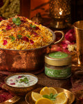 An elaborate close-up shot of a copper pot filled with biryani, a bowl of raita, lemon wedges, and a ZEST Royal Biryani spice blend jar, set on a richly patterned fabric with warm lighting.